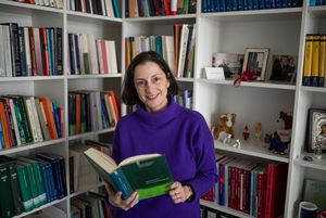 Foto von Artemis Alexiadou. Sie steht mit einem Buch in der Hand vor einem vollen Bücherregal und schaut freundlich lächelnd in die Kamera. 